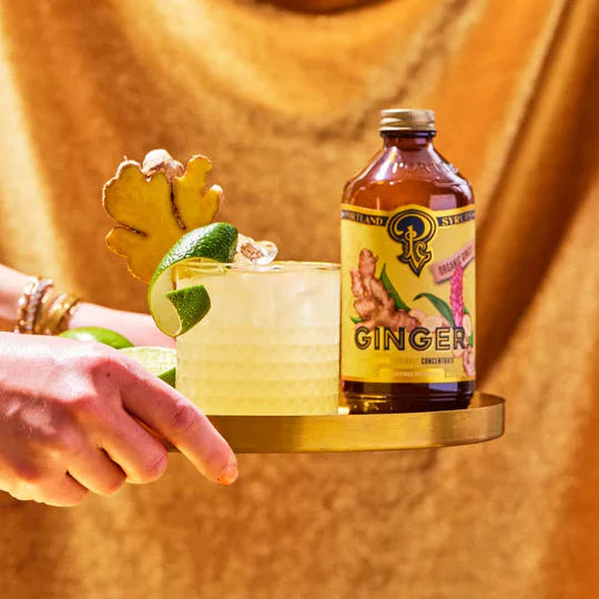 Bottle of ginger ale on a gold tray with a cocktail garnished with lime and ginger root against a draped fabric background.